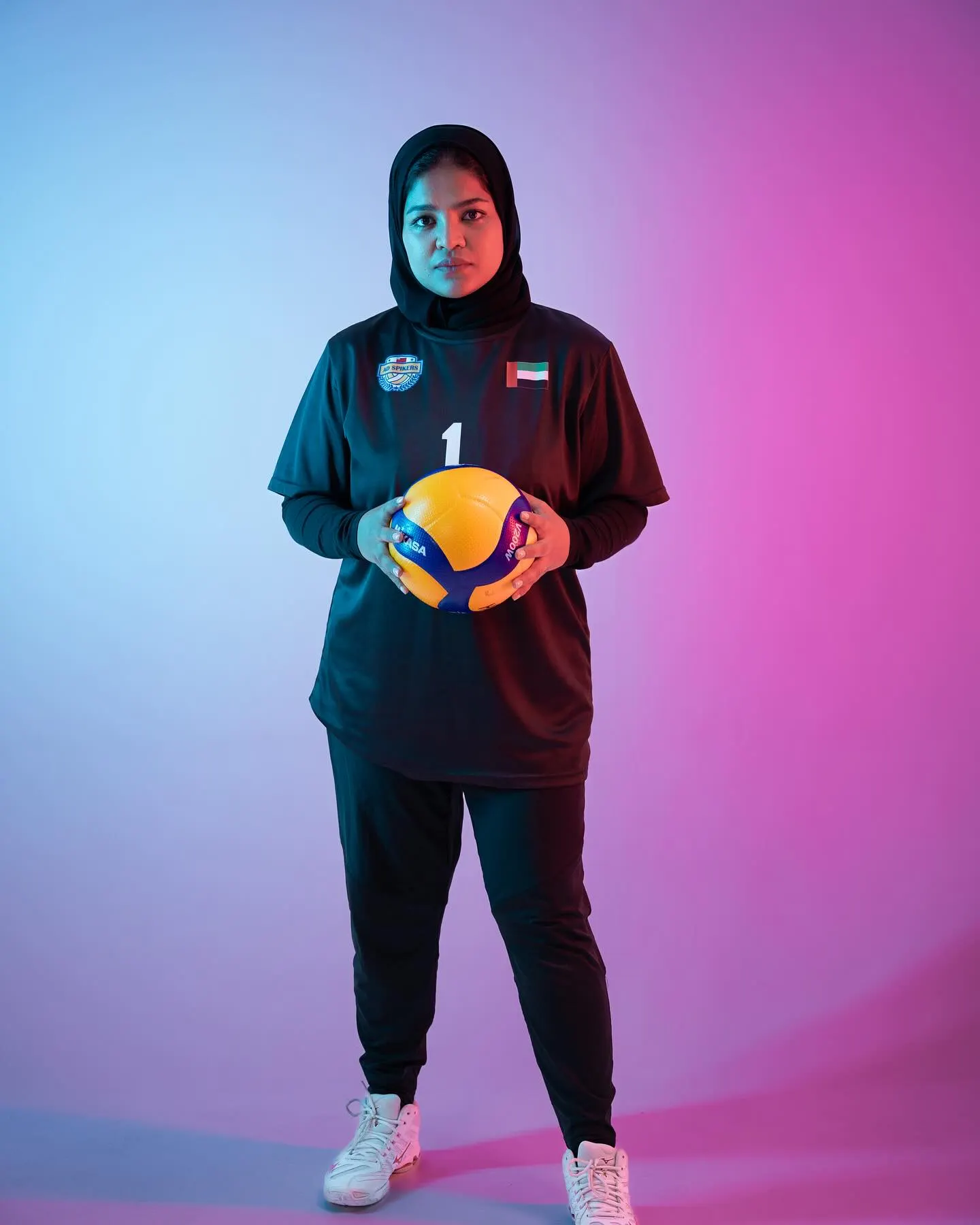 A volleyball player in a dark tracksuit holding a ball, lit against a blue and magenta gradient