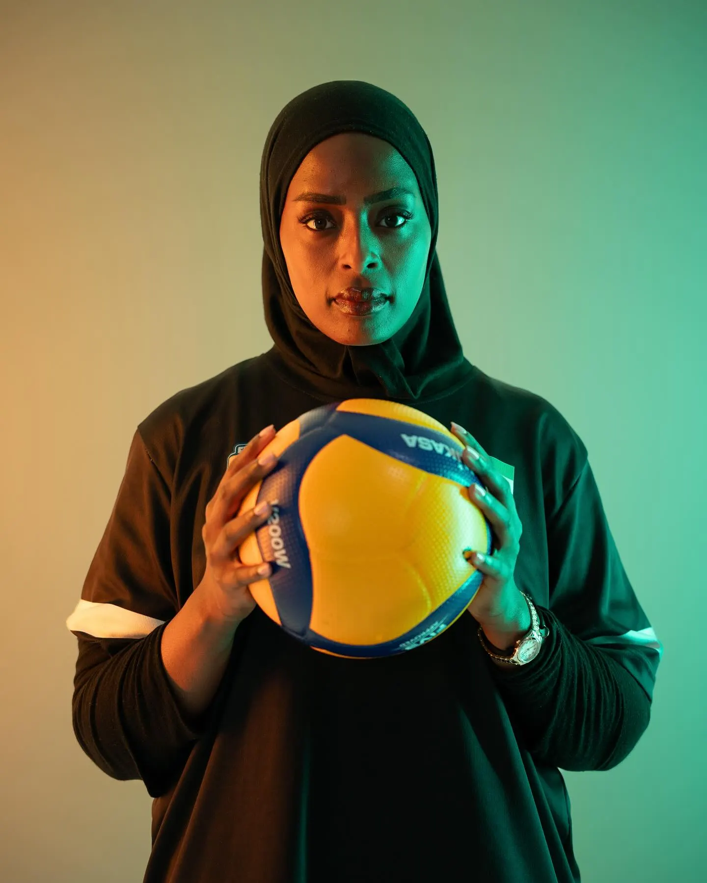 A volleyball player in a dark kit and hijab holding a ball to her chest against a deep green backdrop