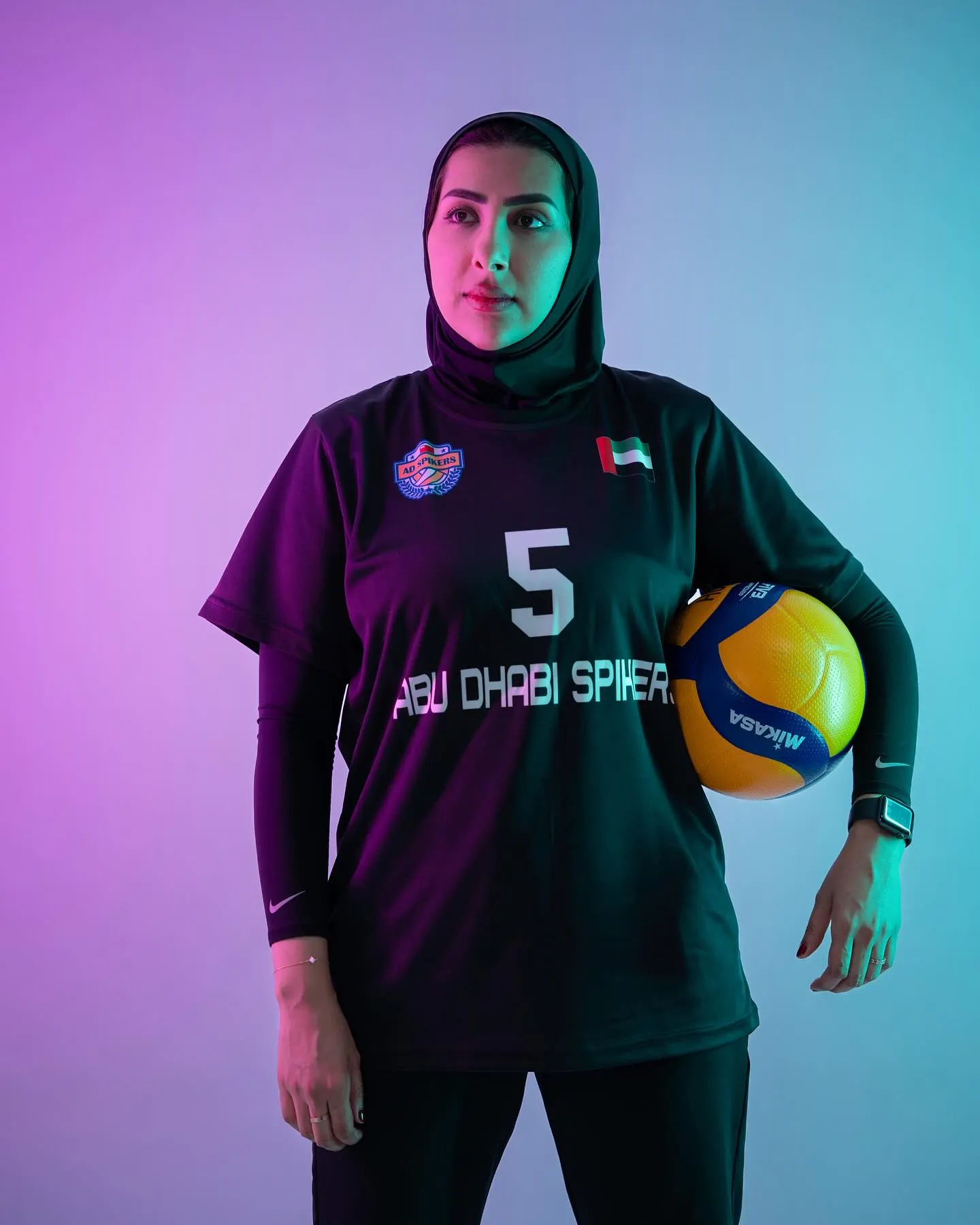 A volleyball player in a dark kit numbered five holding a ball at her hip against a deep green backdrop