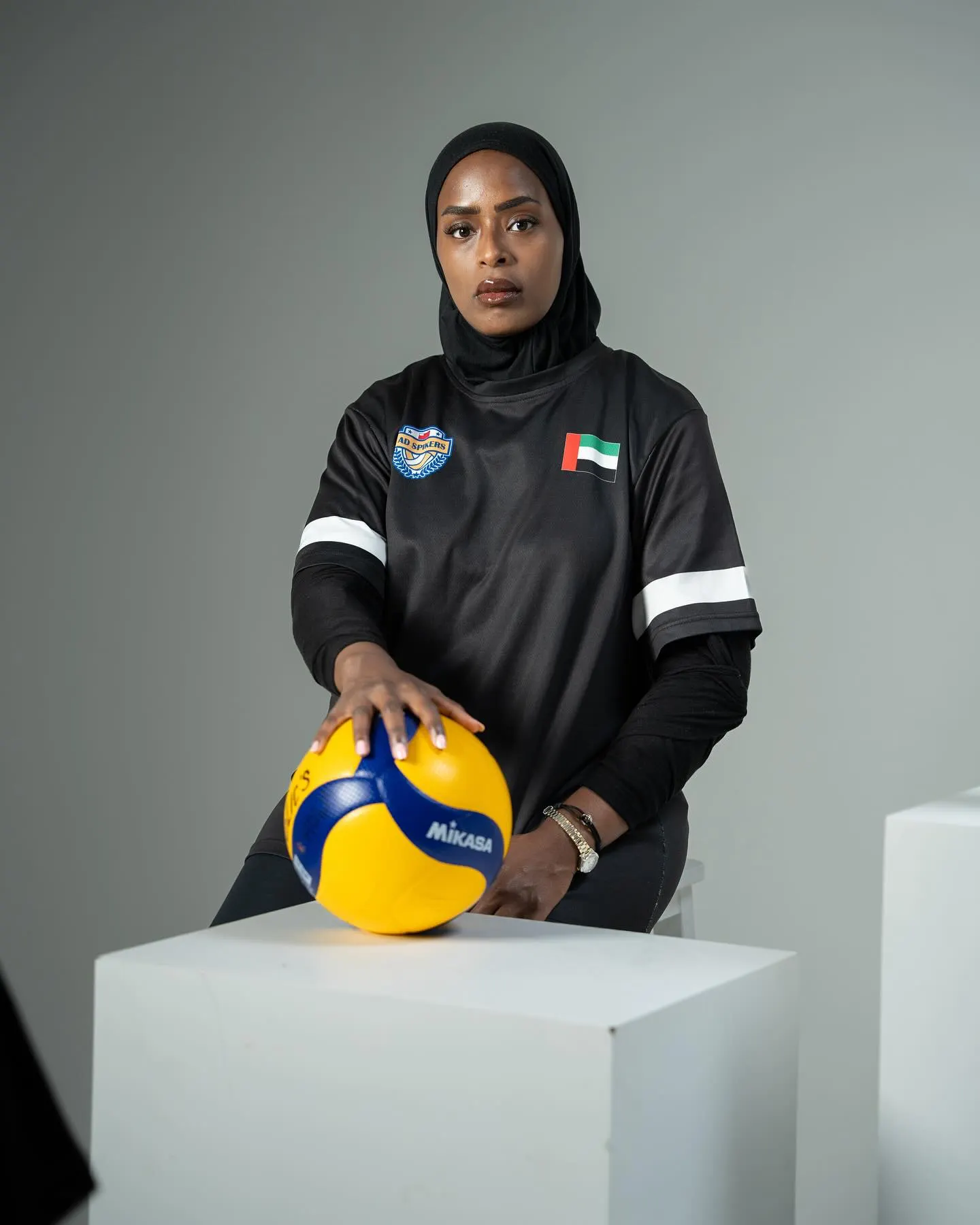 A volleyball player in a dark tracksuit seated on a white plinth holding a ball in her lap