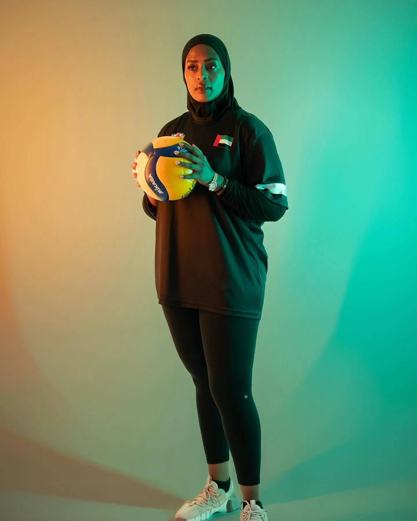 A volleyball player in a dark kit and hijab holding a ball, against a teal and green gradient backdrop