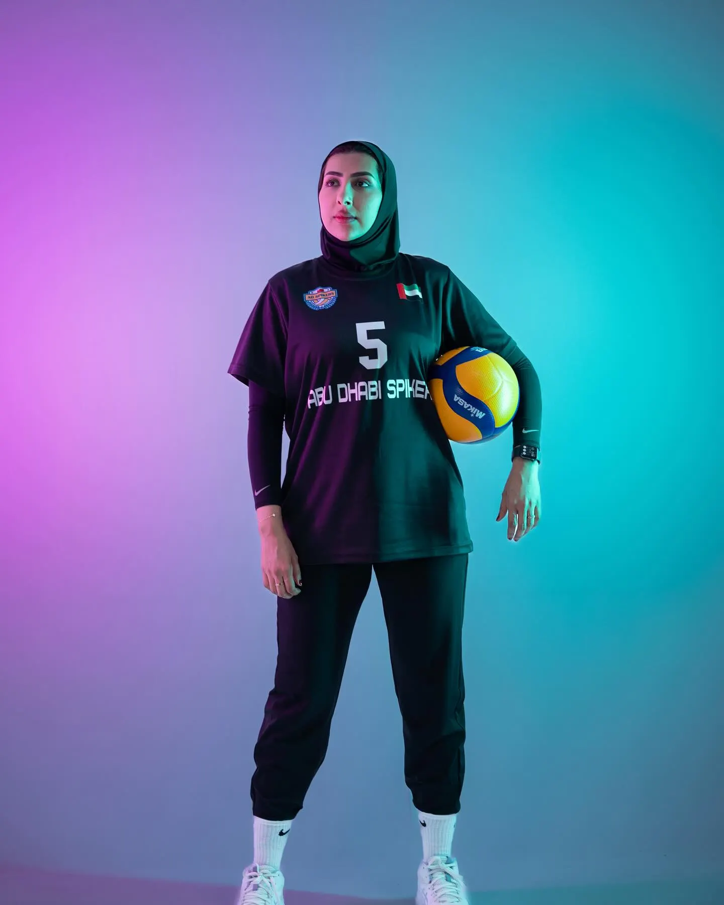 A volleyball player in a dark kit numbered five, holding a ball against a violet and teal gradient backdrop