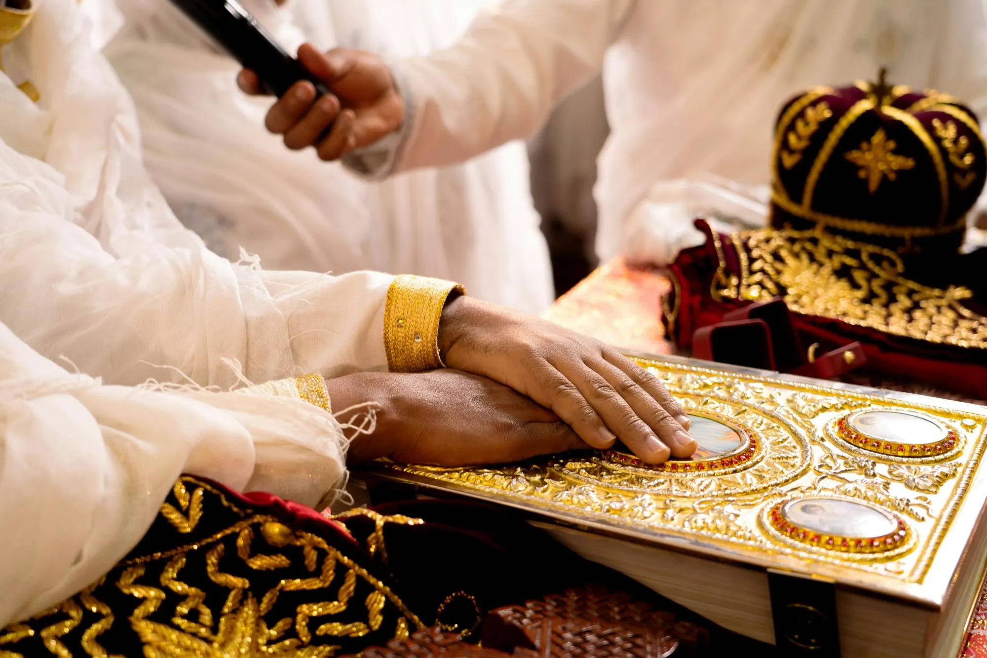 A couple's hands resting together on a gold, jewelled gospel book during a wedding ceremony, with embroidered crowns and robes beside it