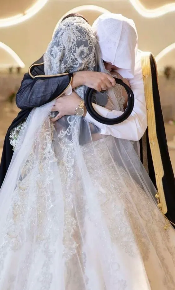 A bride in a beaded cathedral veil and a groom in a black bisht with gold trim holding each other close