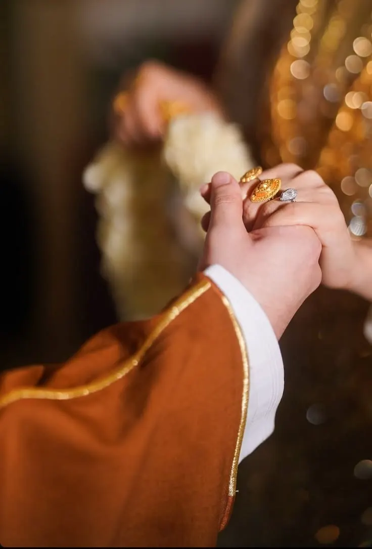 A groom in a rust-coloured bisht with gold trim holding the bride's hand, her fingers wearing gold and diamond rings, against warm golden light