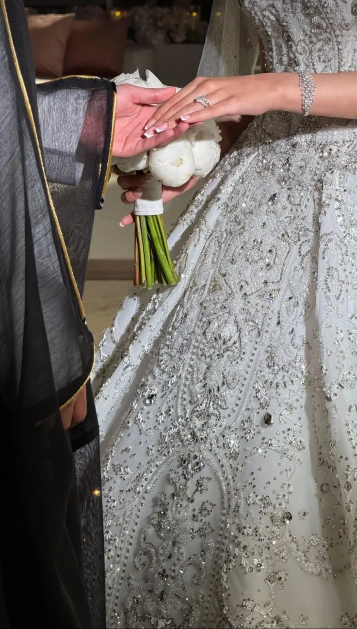 A groom in a black bisht holding the bride's hand, her engagement ring over a bouquet of white peonies and a crystal-beaded gown