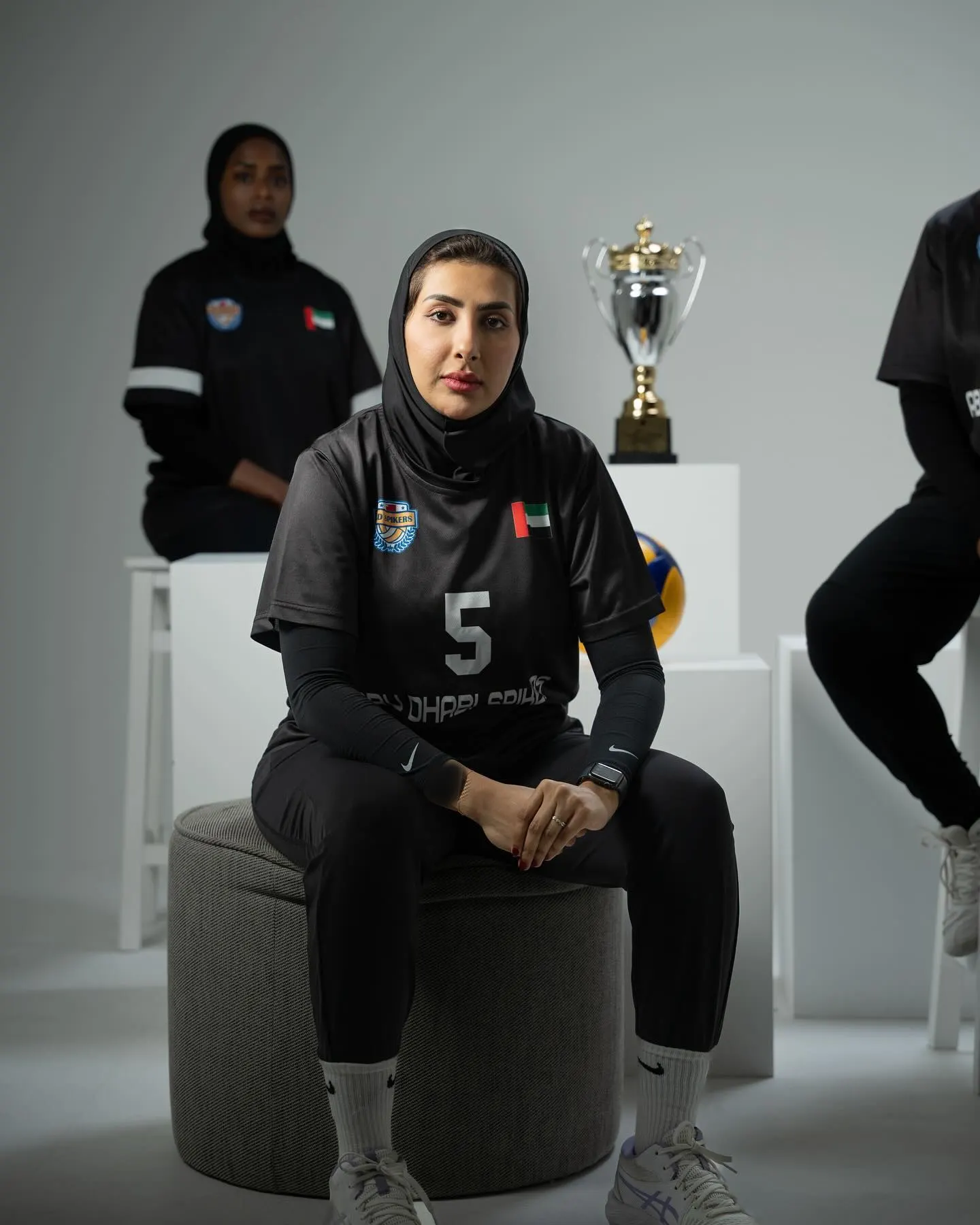 A volleyball player in a black kit and hijab seated facing camera, a trophy and a ball on plinths behind her and two teammates out of focus at the edges of the frame